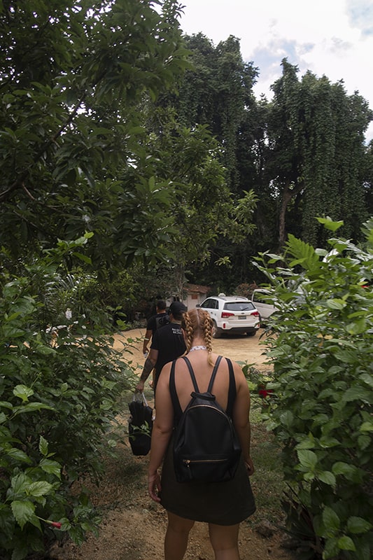a women in braids and a black dress walking through some bushes at the car park in rarru cascades