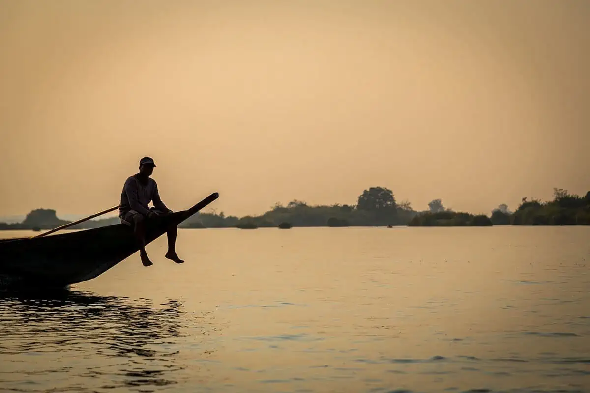a man sitting on the front of a boat in don det during sunset