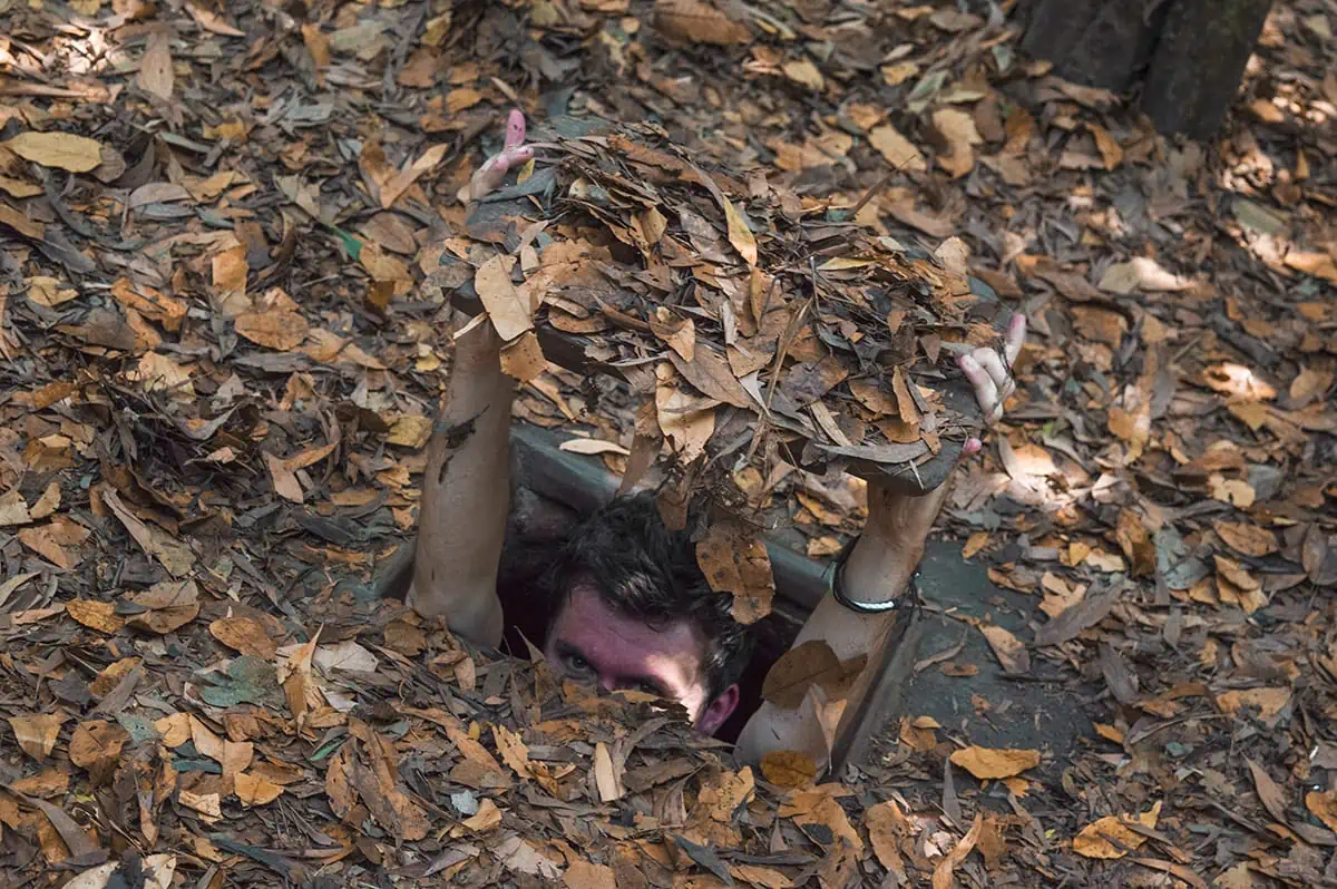 Man emerging from a camouflaged tunnel at cu chi tunnels opening surrounded by fallen leaves, an intriguing historical site for a Vietnam itinerary 3 weeks.