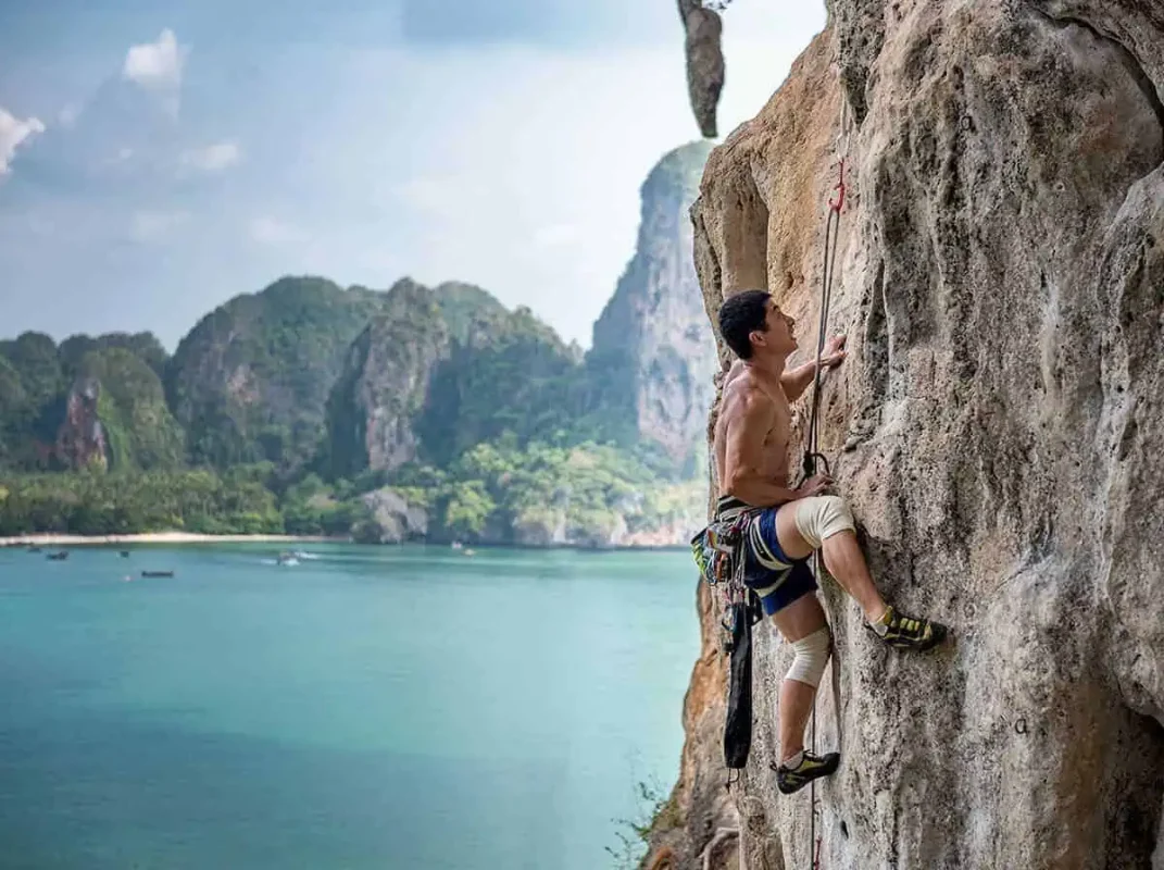 Ao Nang To Railay Beach: The ULTIMATE Guide 2025 13 A rock climber ascending a steep limestone cliff at Railay Beach with a view of the sea and beach behind him.