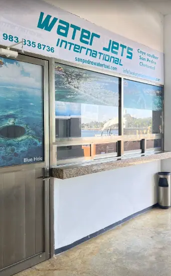 The Water Jets International ticket office, providing ferry services to Caye Caulker, San Pedro, and Chetumal. The office features a sign with contact details and a promotional image of the Blue Hole. 