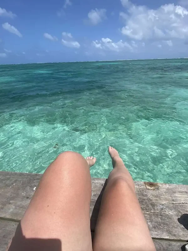 View of a Tasha's legs dangling off a wooden dock, with her feet hovering above the clear waters of Caye Caulker.