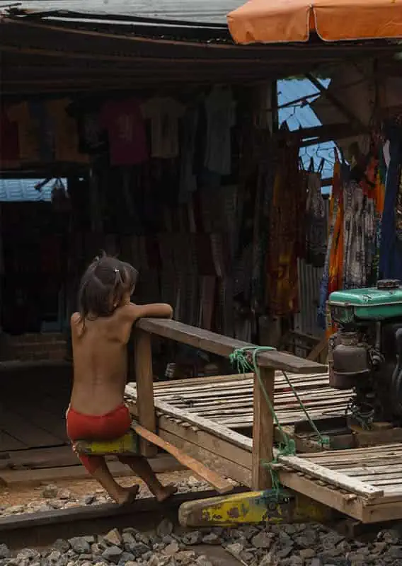 A young child sits at the edge of a bamboo train cart, parked in front of a market stall with hanging clothes and in the background.