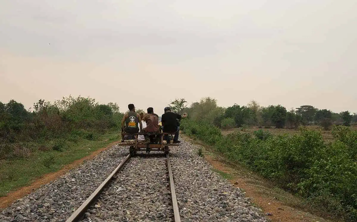 A group of people riding a bamboo train cart along the railway, surrounded by vegetation and open countryside.