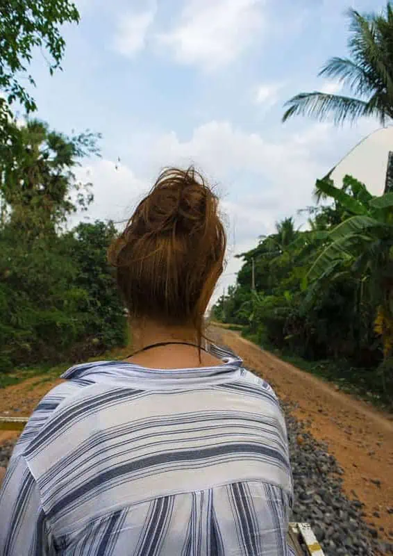 tasha amy with her back to the camera sits on a battambang bamboo train riding along the railway, surrounded by palm trees and jungle.