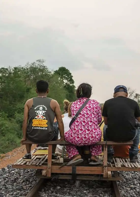 View from behind several locals riding together on a single bamboo train cart, including a man in a tank top with “Cambodia” written on the back.