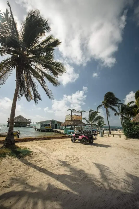 Belize In April: What You NEED To Know 7 A quaint sandy path runs along a serene beach in Belize, flanked by rustic buildings and thatched-roof bars. A pink golf cart parked on the side adds a touch of modern convenience to this otherwise natural and tranquil setting.