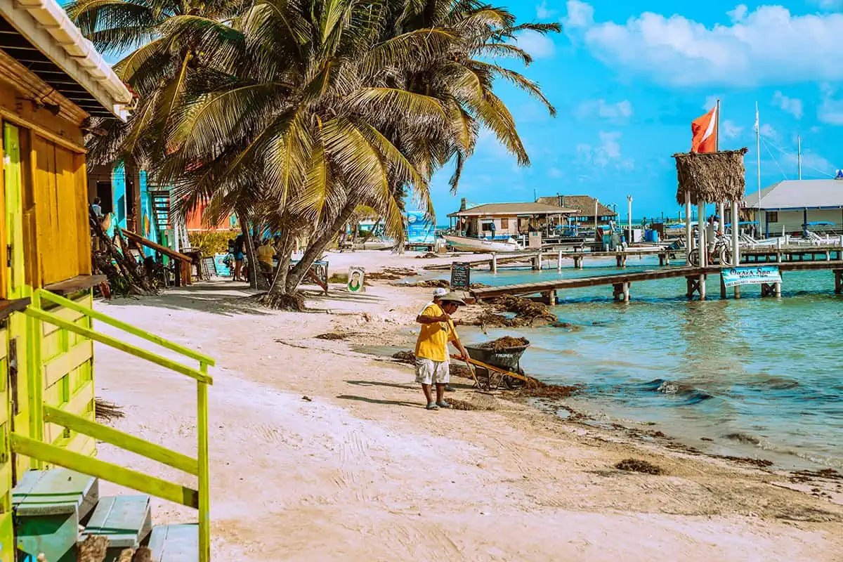 Belize In June: Is Belize Worth Visiting In June? 12 A local Belizean in a yellow shirt walking along a beachside path with wooden docks and colorful buildings, typical of a June day in Belize.