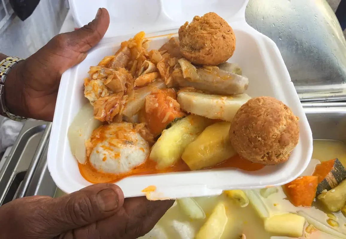 Hand holding a Styrofoam container filled with a Jamaican Ackee and Saltfish breakfast, complete with johnny cakes and root vegetables, ready for a morning on the go.