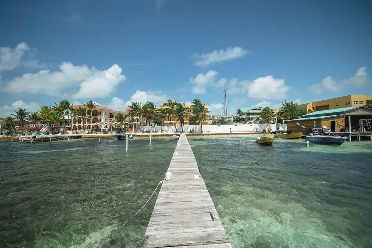 A wooden pier stretching into the clear green-blue waters with a view of the beachfront and palm trees, illustrating the tranquil and clear waters that make Ambergris Caye a top beach destination.