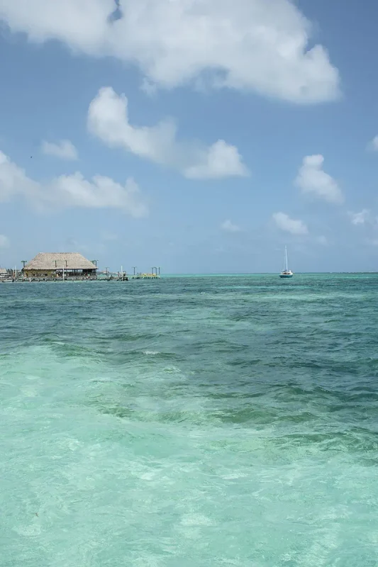 A view of a calm sea with a thatched hut on a pier and a sailboat in the distance under a vast blue sky, highlighting the serene beauty of Ambergris Caye's beaches.