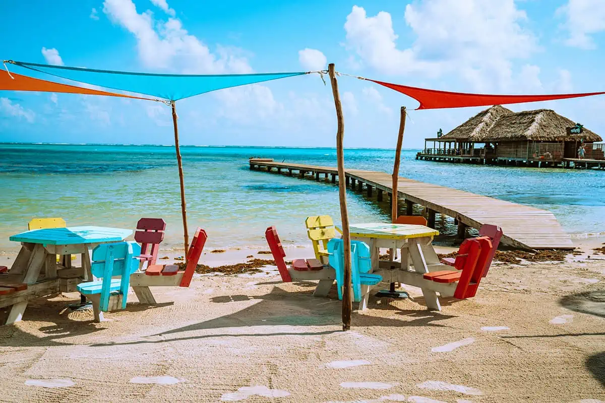 Colorful beach chairs and a picnic table under shade sails on the sandy shores of Ambergris Caye, with a view of the calm turquoise waters and a thatched pier in the background, inviting relaxation at one of the best beaches.