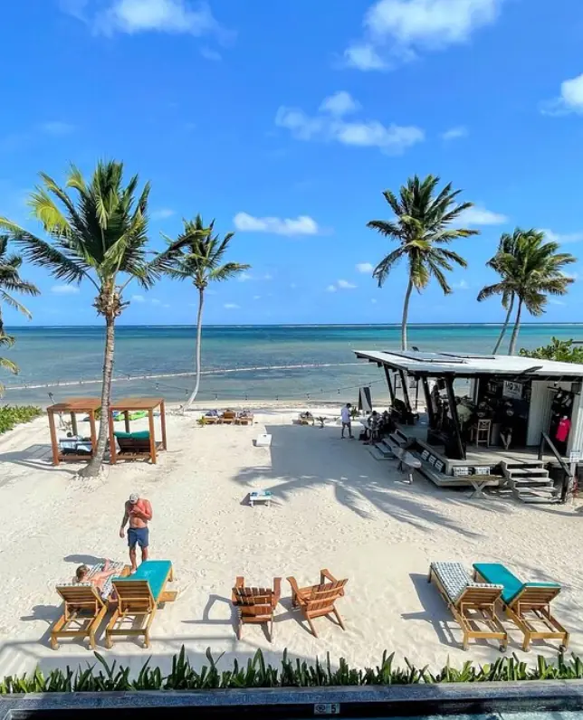 Elevated view of a beachfront with loungers and palm trees, overlooking a tranquil sea and a beach bar, capturing a typical leisurely day on Ambergris Caye.