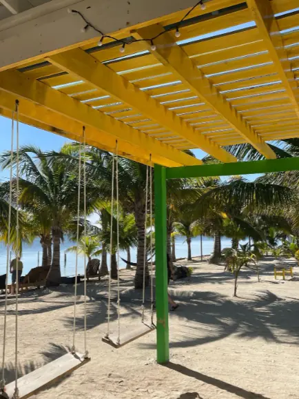 A view under a bright yellow pergola with swings, looking out to a sandy beach with palm trees and clear skies, a relaxing corner on Ambergris Caye.