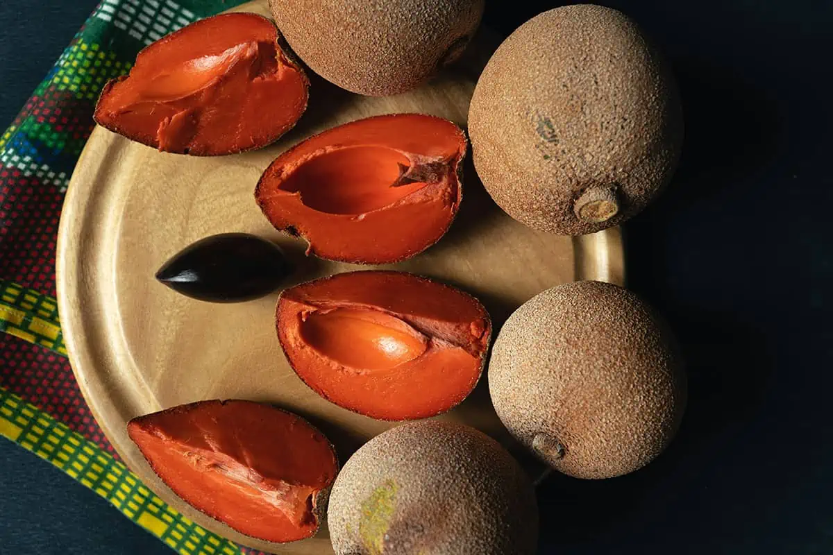 A wooden plate displaying open mamey sapote fruits with reddish-orange flesh and a glossy black seed, a tropical fruit often enjoyed in Belize.