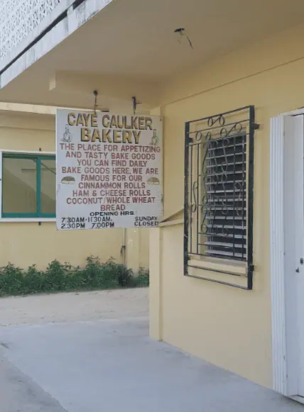 Exterior view of Caye Caulker Bakery, prominently featured as one of the best restaurants on Caye Caulker. The bakery's facade is painted a warm yellow, and a large white sign hangs above a barred window, detailing the delicious offerings such as daily fresh cinnamon rolls and whole wheat bread. This bakery is highlighted as the place for 'appetizing and tasty bake goods' in Caye Caulker, open daily with posted hours for convenient visitor access.