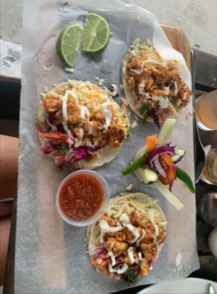 Close-up of three shrimp tacos served on a paper-lined tray at Barrier Reef Sports Bar & Grill, one of the best restaurants on Caye Caulker. Each taco is filled with crispy fried shrimp, fresh cabbage slaw, and drizzled with creamy sauce, accompanied by a lime wedge and a small bowl of salsa, providing a colorful and appetizing presentation