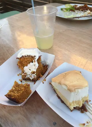 A casual dining setting at Chef Juan's Kitchen and Pastries featuring two popular desserts: a slice of carrot cake topped with cream cheese frosting and a slice of creamy pie, both served on paper plates. A cup of lemonade sits nearby, complementing the sweet treats, indicative of the relaxed yet indulgent dining experience at this Caye Caulker spot.