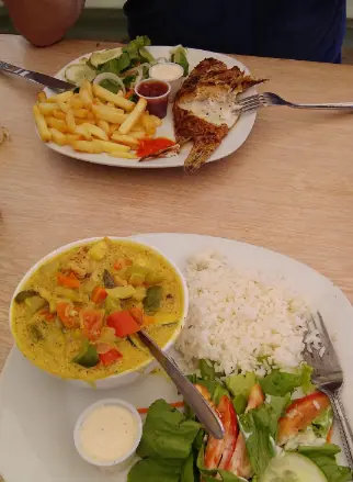 A table at Chef Juan's Kitchen and Pastries, renowned as one of the best restaurants on Caye Caulker, showcasing two plates of hearty meals. The first plate features grilled fish with a side of fries and a garden salad, and the second plate offers a curry dish with mixed vegetables served alongside white rice and a green salad, highlighting the fresh, local ingredients used in their cuisine.