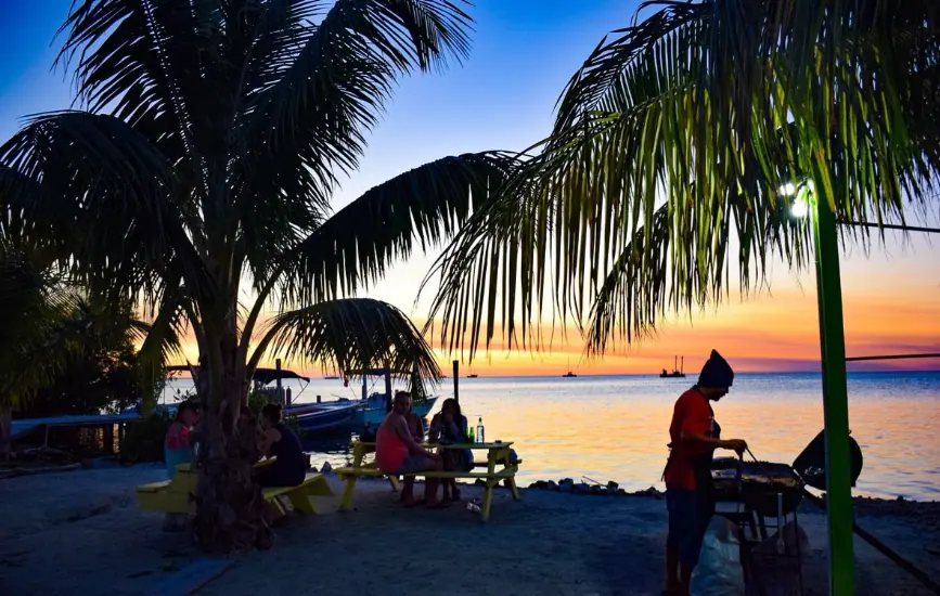 An enchanting evening scene at Dee N D's Waterside Eatery, widely regarded as one of the best restaurants on Caye Caulker. The photograph captures diners seated at bright yellow picnic tables under the swaying palm trees along the beachfront. A cook in a red shirt is barbecuing by the shore as the sun sets in the background, painting the sky in vivid hues of orange and blue, reflecting off the calm Caribbean waters. This setting exemplifies a perfect tropical dining experience on Caye Caulker.