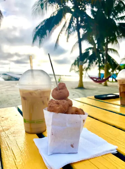 A delightful morning snack scene at Ice N Beans, one of the best restaurants on Caye Caulker for a quick bite. A paper cone filled with freshly made donuts, dusted with cinnamon sugar, is paired with a tall plastic cup of iced coffee on a yellow picnic table. The table is set against a backdrop of a sandy beach and palm trees, offering a quintessential tropical setting.