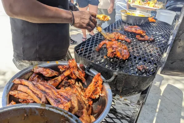 A bustling street food scene at Chef Kareem's UnBelizeable Lunch, known as one of the best restaurants on Caye Caulker for authentic local cuisine. The photo shows a chef, deftly handling various pieces of juicy, barbecued chicken and ribs over a grill, with a bowl of marinade at hand for basting. The grill is filled with deliciously charred meats, enticing passersby with its smoky aroma. This image captures the vibrant street food culture that is a hallmark of Caye Caulker’s culinary offerings.
