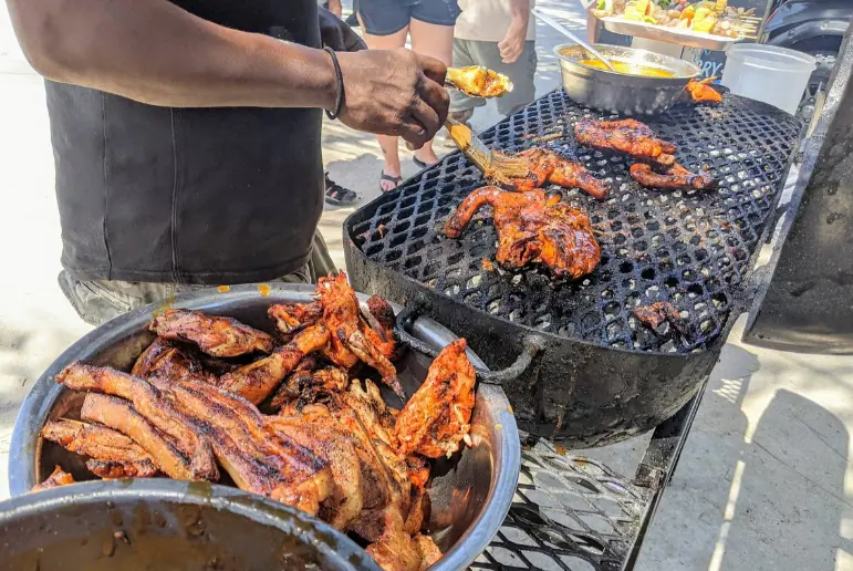 A bustling street food scene at Chef Kareem's UnBelizeable Lunch, known as one of the best restaurants on Caye Caulker for authentic local cuisine. The photo shows a chef, deftly handling various pieces of juicy, barbecued chicken and ribs over a grill, with a bowl of marinade at hand for basting. The grill is filled with deliciously charred meats, enticing passersby with its smoky aroma. This image captures the vibrant street food culture that is a hallmark of Caye Caulker’s culinary offerings.