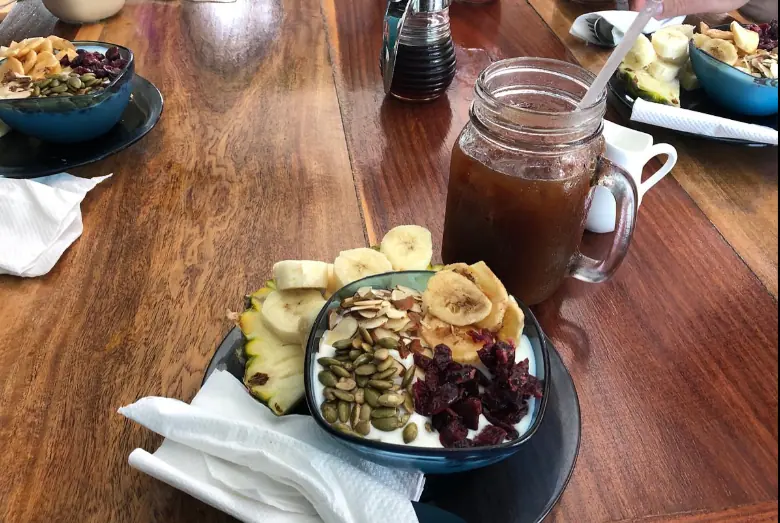 A healthy and vibrant breakfast spread at Namaste Cafe, renowned as one of the best restaurants on Caye Caulker for healthy dining. The image displays a bowl of yogurt topped with sliced bananas, pineapple, pumpkin seeds, and dried cranberries. Adjacent to it is a mason jar filled with iced tea, reflecting a refreshing start to a day. The meal is set on a polished wooden table, adding warmth to the wholesome and inviting atmosphere of the cafe