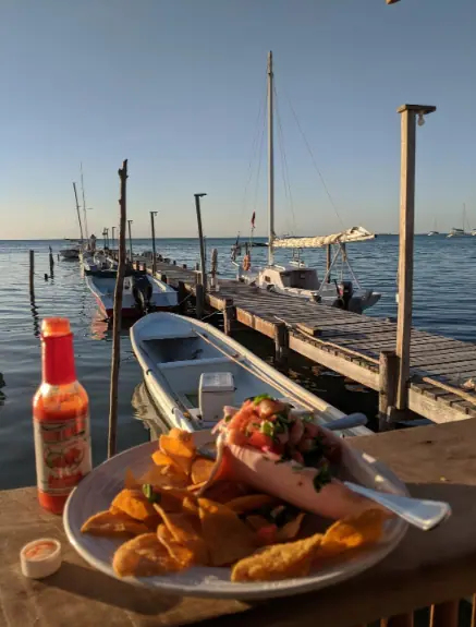 A picturesque dining experience at Pelican Sunset Bar, one of the best restaurants on Caye Caulker, featuring a plate of shrimp cocktail and fried plantains. The dish is served on a rustic wooden table overlooking a pier with moored boats and the calm Caribbean Sea, capturing the essence of a perfect seaside meal at sunset