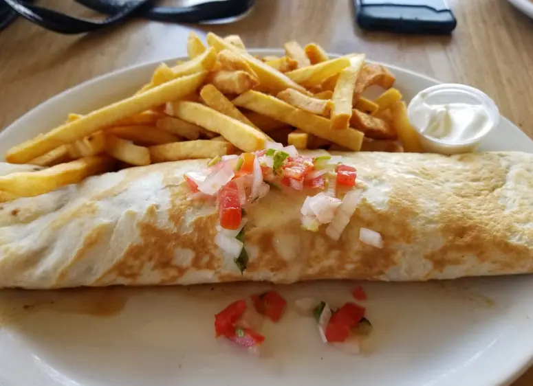 A classic lunch serving at Rainbow Grill & Bar, popularly known as one of the best restaurants on Caye Caulker. The plate features a large, golden-brown wrap topped with fresh pico de gallo, served alongside a generous portion of crispy French fries and a side of sour cream. This dish is presented on a simple white plate, symbolizing the straightforward yet delicious offerings of the restaurant, ideal for a satisfying meal after exploring the island