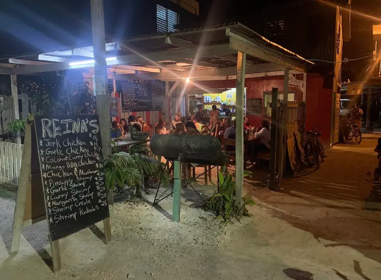 A lively evening scene at Reina's, a popular outdoor dining spot on Caye Caulker. The image captures a rustic, open-air restaurant bustling with diners seated at simple wooden tables. A chalkboard menu stands at the entrance, listing an array of dishes such as Jerk Chicken and Coconut Curry Chicken, illuminated by overhead string lights that add a cozy glow to the setting. A large, barrel-style grill is active in the background, with a chef preparing meals, enhancing the casual, communal atmosphere of the restaurant
