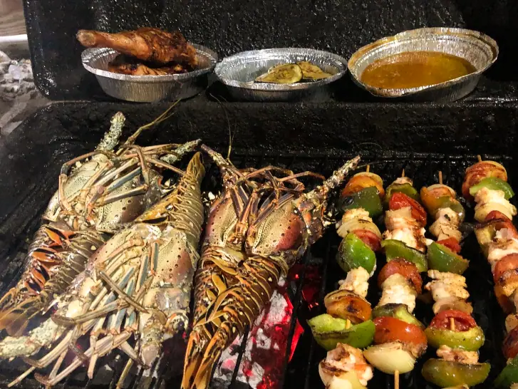A vibrant grill scene at Steve's Grill, showcasing a variety of freshly grilled seafood and meats. The image features spiny lobsters and skewers of chicken, sausage, bell peppers, and onions sizzling over a charcoal grill. To the side, there are two aluminum foil containers: one with grilled plantains and the other with a golden, savory sauce. The lively, crackling fire underlines the freshness and local flavor of the seafood, offering a glimpse into the casual, flavorful dining experience at the grill.