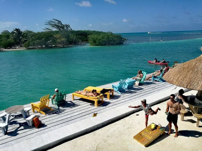 A lively scene at a waterfront area in Caye Caulker, with people lounging on colorful Adirondack chairs and sunbeds along a wooden deck. Others are engaged in outdoor activities, with kayaks and a small boat visible on the calm, turquoise water. This vibrant atmosphere showcases one of the best things to do in Caye Caulker—relaxing and enjoying the waterfront activities.