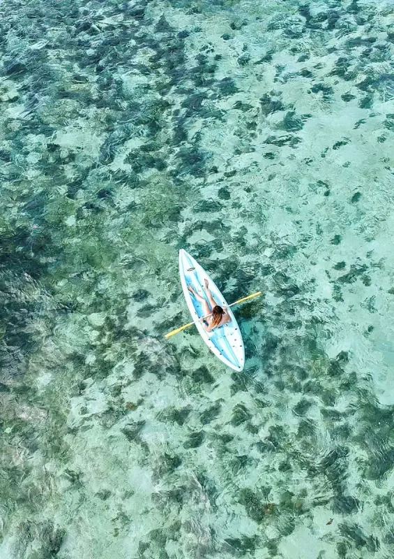 Aerial view of a woman kayaking on the clear, shallow waters of Caye Caulker. The water's transparency reveals the seagrass below, creating a stunning mosaic of blues and greens. Kayaking is one of the best things to do in Caye Caulker, offering a peaceful way to explore the marine environment.