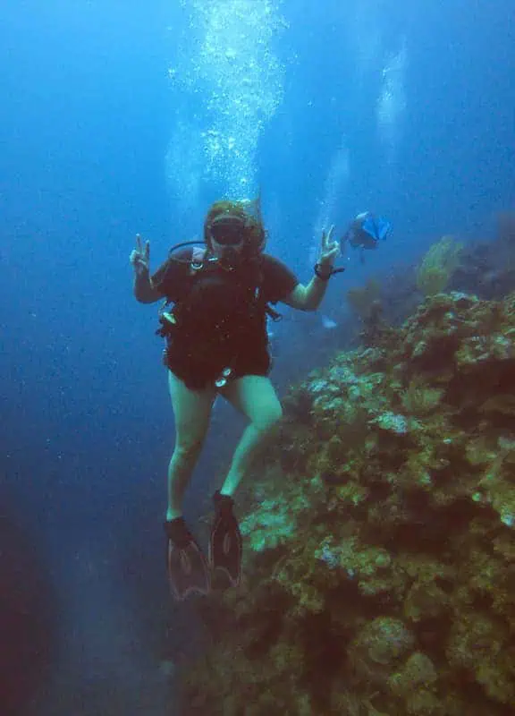 A scuba diver giving a peace sign while exploring a coral reef in the deep blue waters off Caye Caulker. The diver is surrounded by marine life and colorful coral formations, showcasing the vibrant underwater ecosystem.