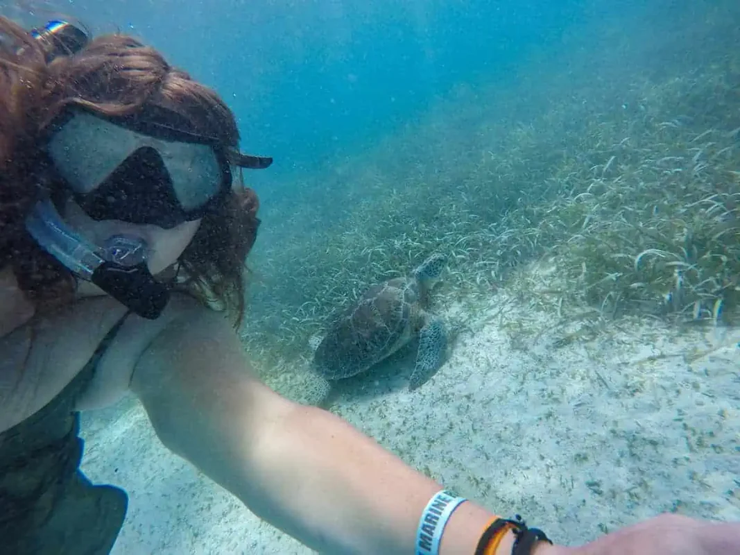 tasha amy captures a selfie with a sea turtle swimming just behind her in the shallow, clear waters of Caye Caulker. The snorkeler wears a mask and snorkel, her hair floating around her as she takes the photo. This close encounter with a sea turtle, one of the majestic creatures of the ocean, is among the best things to do in Caye Caulker.