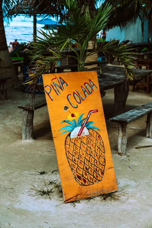 A colorful wooden sign painted with a large pineapple and the words "Piña Colada," placed on sandy ground near rustic picnic tables and palm trees. This sign, near a beach bar, invites visitors to enjoy a refreshing tropical drink.