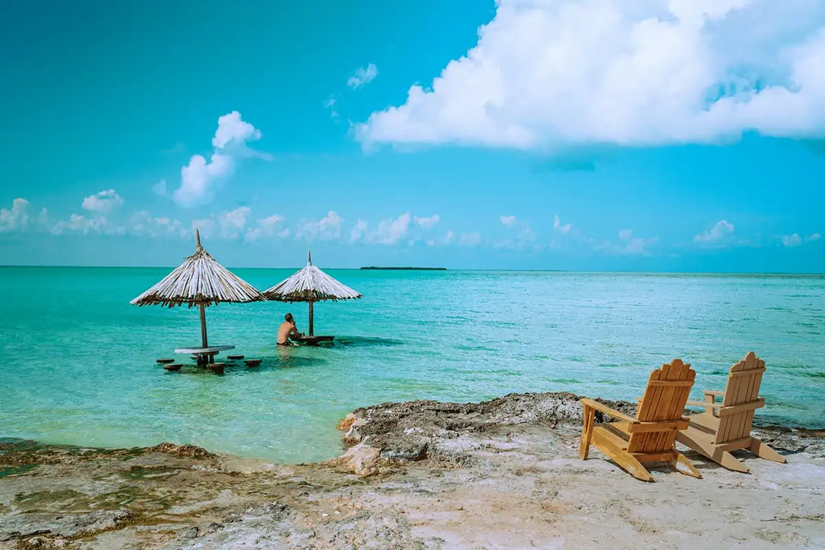 A tranquil beach scene in with two thatched-roof parasols and wooden picnic tables partially submerged in the clear turquoise water. A person is seen enjoying the water while two wooden chairs sit on the rocky shore, offering a perfect spot to relax. This serene setup highlights one of the best things to do in Caye Caulker—going island hopping