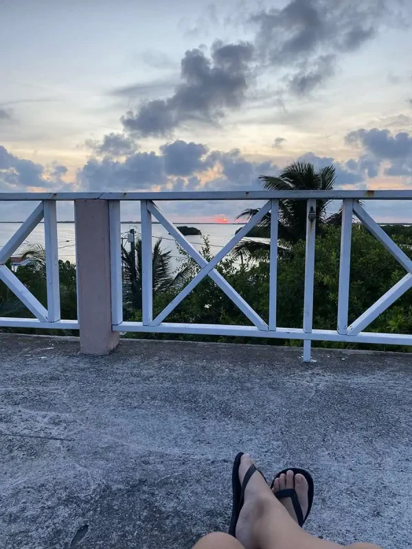 A tranquil scene at dusk on Ambergris Caye with a person's legs resting on a balcony railing, showcasing their relaxed posture and flip-flops with a stunning view of the ocean and a vibrant sunset in the background.