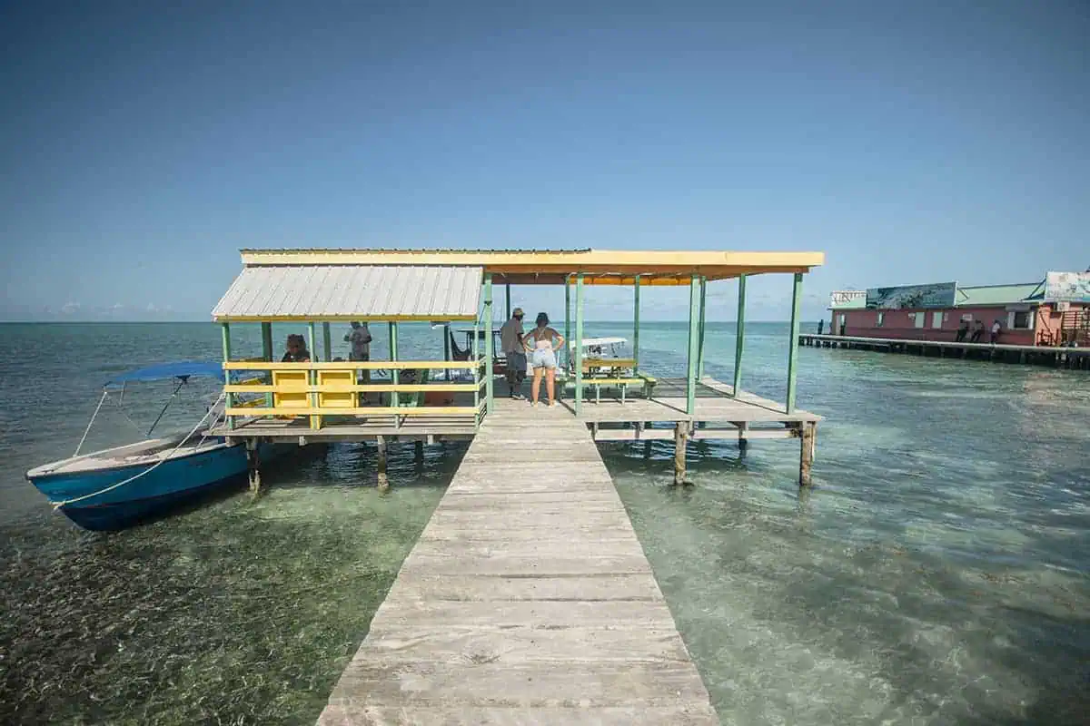 A rustic wooden dock in Caye Caulker extending into the turquoise sea with a boat tied up and people enjoying the view, a common relaxing activity on the island.