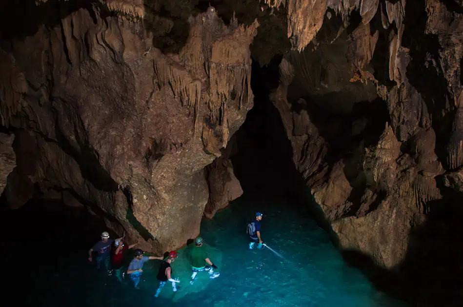Group of people wading through crystal-clear water inside a dark, ATM cave. They are wearing helmets and headlamps, and the scene captures the sense of adventure and excitement, highlighting one of the thrilling things to do in San Ignacio.