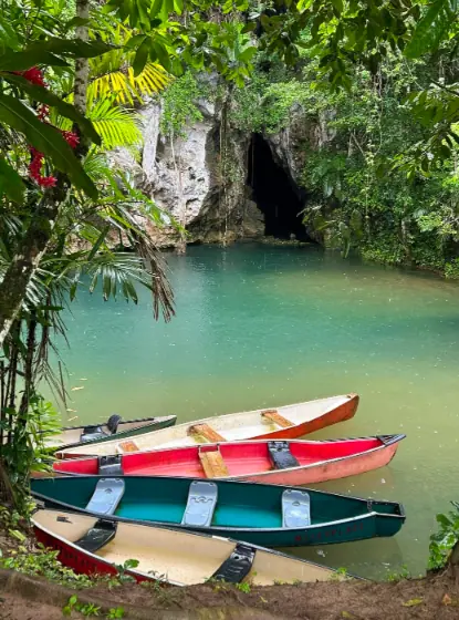 Colorful canoes docked by the edge of a serene, turquoise river. The river is surrounded by lush tropical vegetation, and the barton creek cave entrance is visible in the background, offering a picturesque and peaceful setting for outdoor activities in San Ignacio.
