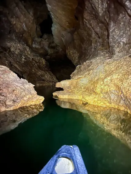 Inside a narrow cave passage with clear, emerald-green water. The rocky walls of the cave are illuminated, showcasing the textures and formations, and emphasizing the exciting caving and exploration opportunities available in San Ignacio