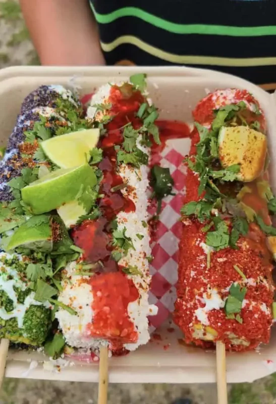 Close-up of a tray with three colorful, seasoned corn on the cobs, garnished with lime wedges and herbs. The vibrant presentation of the food highlights the rich culinary experiences available in San Ignacio