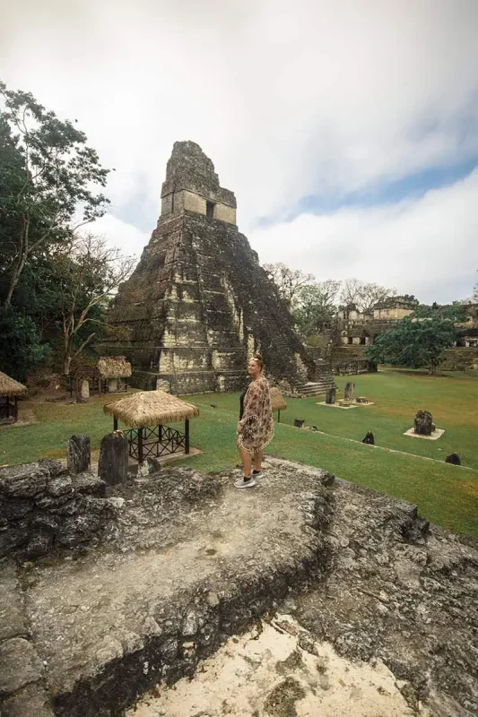 tasha amy standing in front of a towering Mayan pyramid at Tikal, surrounded by lush greenery and other ancient structures. The pyramid's steep steps and weathered stone highlight the historical significance and cultural heritage of this site in San Ignacio