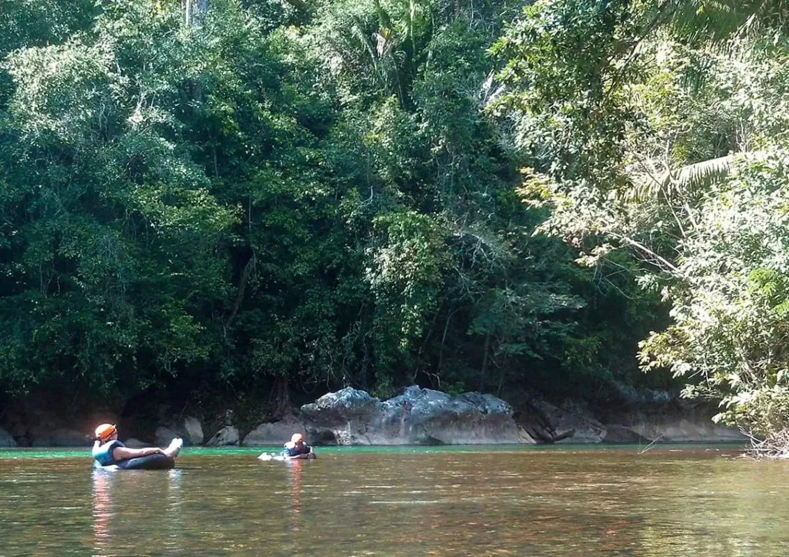 Two people floating on inner tubes in a calm river surrounded by dense jungle in San Ignacio. The participants are wearing life vests and helmets, enjoying the peaceful tubing experience as one of the adventurous things to do in San Ignacio.