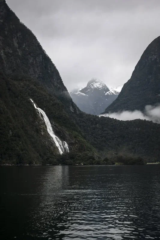 A close-up view of Lakes Bowen Falls in Milford Sound, with the waterfall dramatically descending between two steep, forested cliffs. Snow-capped peaks loom in the background, partially obscured by mist and low clouds. The calm, reflective water below captures the stark contrast of the bright waterfall against the dark, verdant landscape.