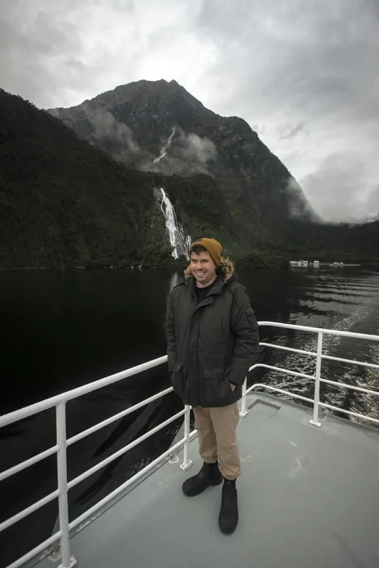 A person in a dark green parka with a fur-lined hood and a mustard yellow beanie stands on a boat deck in Milford Sound. The individual smiles as they pose against the railing, with the majestic Bowen Falls in the background, cascading down a steep, forested mountain. The overcast sky and the dark, reflective water enhance the serene and dramatic atmosphere of the scene.