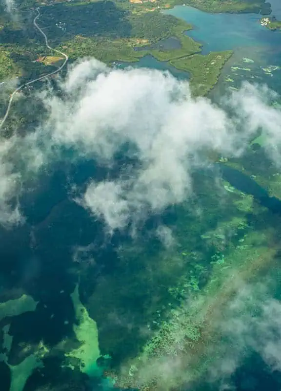 A stunning aerial view from a plane window revealing the vibrant turquoise waters of the Caribbean Sea surrounding the coastline of Caye Caulker, Belize. The foreground shows the plane's metallic railing, while below, dense mangrove forests, a winding road, and a sandy shoreline characterize the island. This captivating sight greets travelers flying from Cancun to Caye Caulker, offering a glimpse of the paradise below