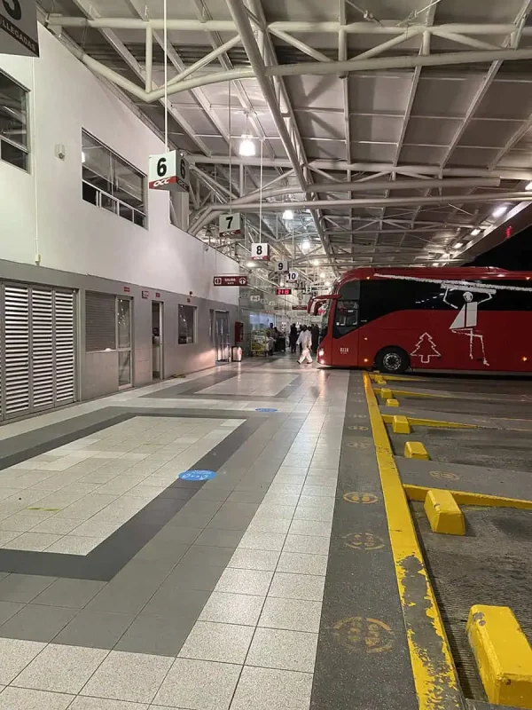 A large, brightly colored red bus parked at a bus terminal in Cancun, Mexico. The terminal features numbered signs and a modern roof structure, with passengers and staff moving in the background. Cancun to Caye Caulker travelers often start their journey from this terminal.
